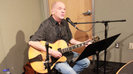 An adult sitting and playing an acoustic guitar and singing into a microphone in a radio studio