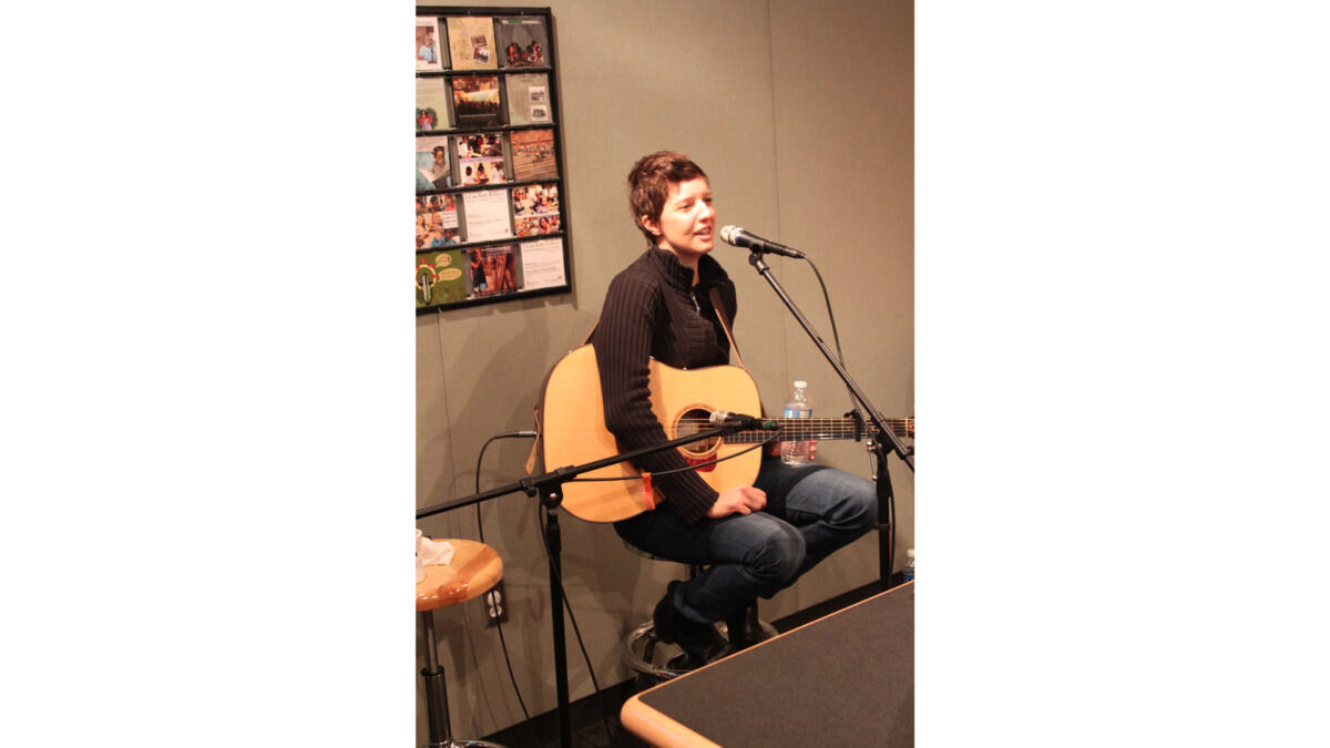 An adult sitting and holding an acoustic guitar while singing into a microphone in a radio studio