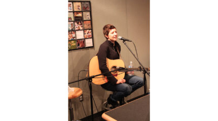 An adult sitting and holding an acoustic guitar while singing into a microphone in a radio studio