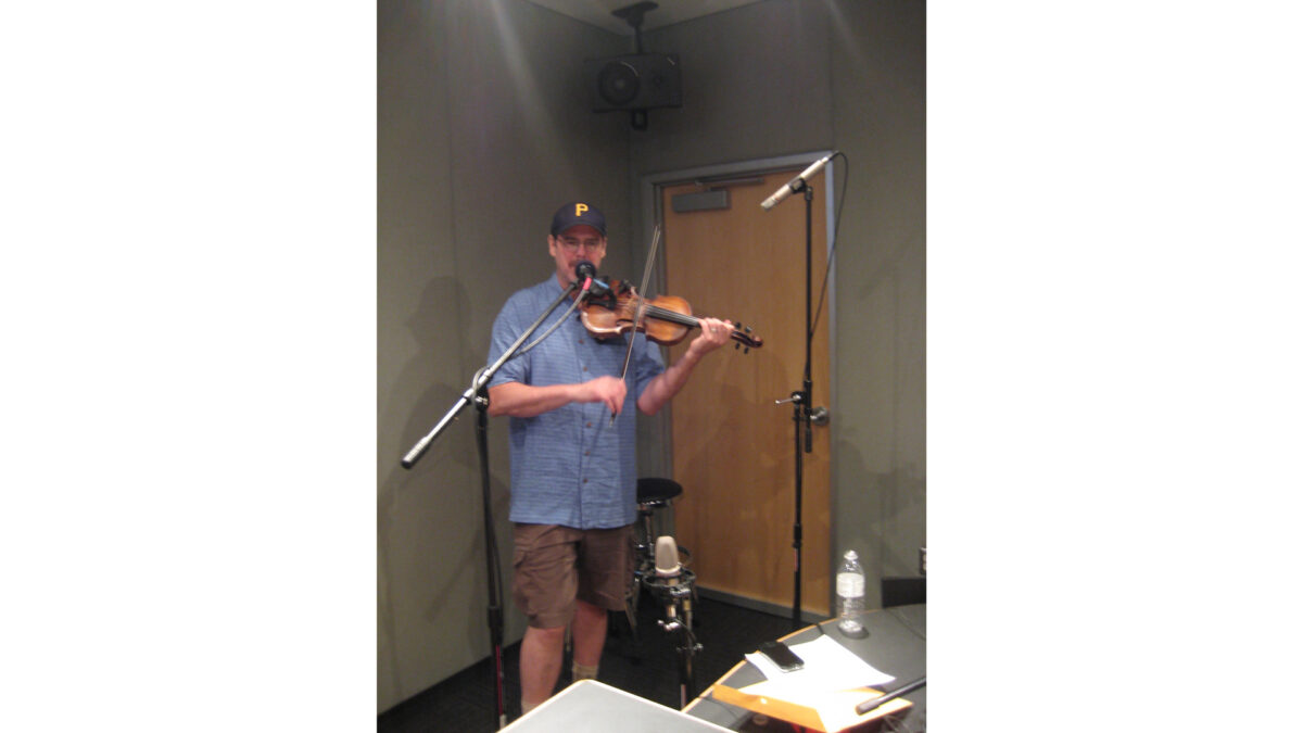An adult playing a violin and singing into a microphone in a radio studio