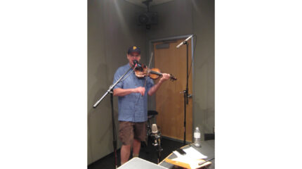 An adult playing a violin and singing into a microphone in a radio studio