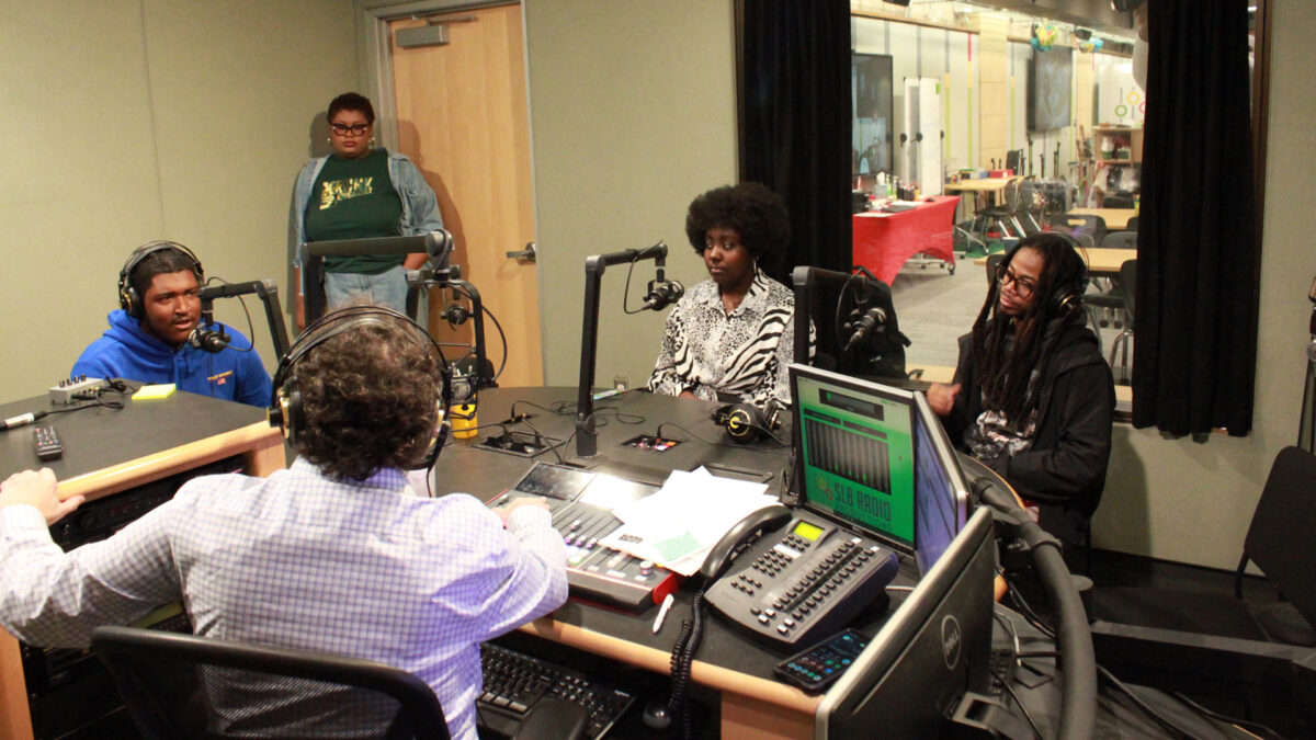 Two teens and an adult speaking into microphones being interviewed by an adult wearing headphones sitting behind a microphone and an audio mixer and an adult standing in the background in a radio studio