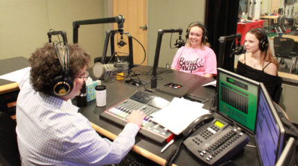 Two teens wearing headphones smiling while sitting behind microphones being interviewed by an adult wearing headphones sitting behind a microphone and an audio mixer in a radio studio