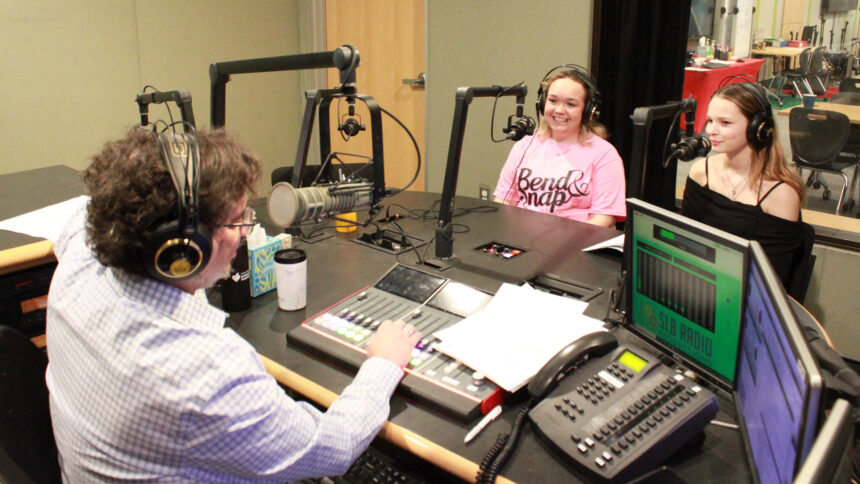 Two teens wearing headphones smiling while sitting behind microphones being interviewed by an adult wearing headphones sitting behind a microphone and an audio mixer in a radio studio
