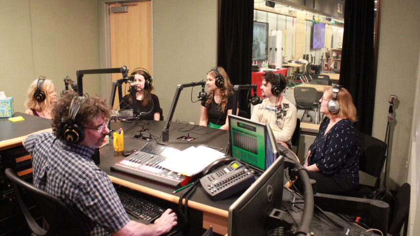 Three teens and two adults wearing headphones sitting behind microphones being interviewed by an adult wearing headphones sitting behind a microphone and an audio mixer in a radio studio