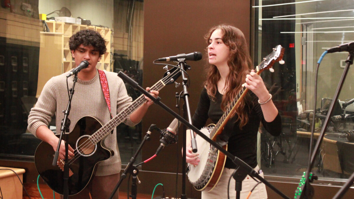 A young adult playing a bass guitar and singing into a microphone and a young adult playing banjo and singing into a microphone