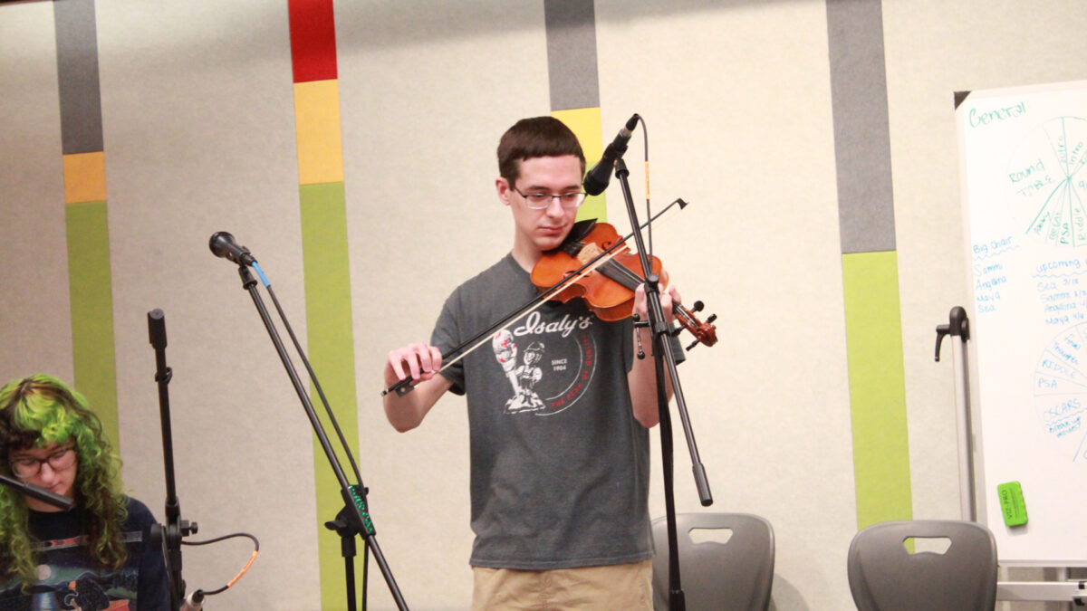 A young adult playing a violin while standing behind a microphone and a young adult sitting behind a microphone