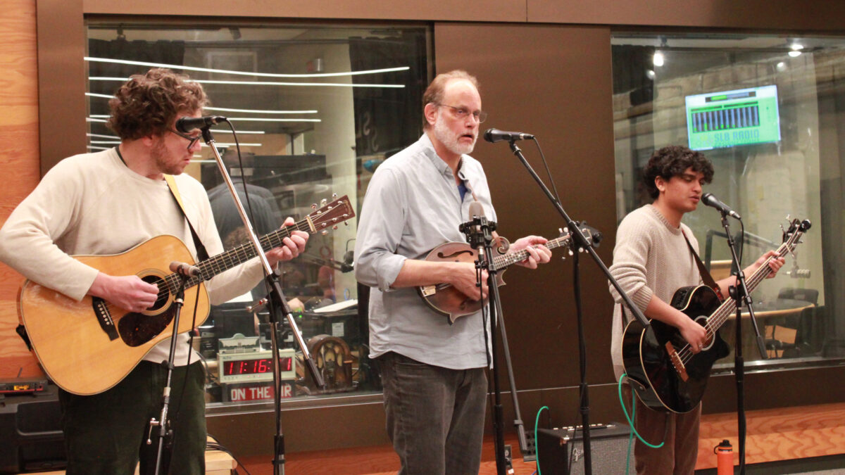 An adult playing an acoustic guitar and standing behind a microphone, an adult playing a mandolin and standing behind a microphone, and a young adult playing a bass guitar and singing into a microphone