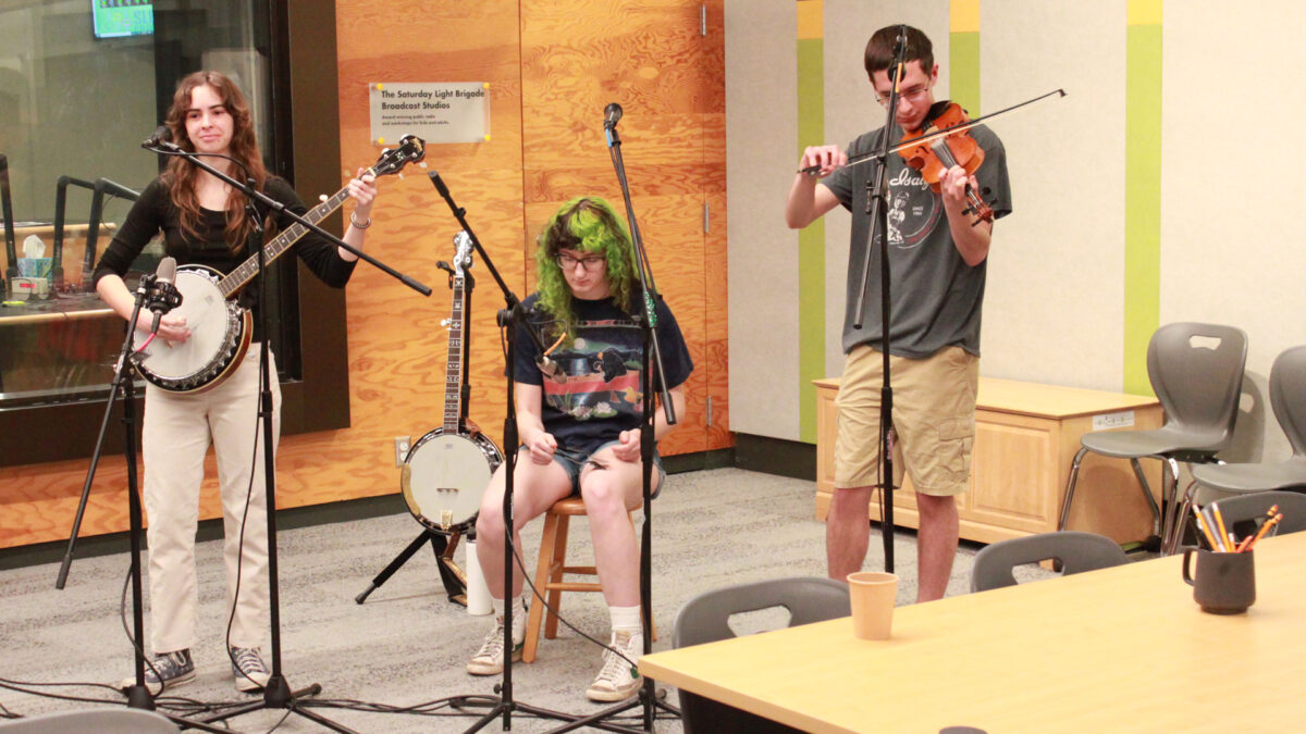 A young adult playing an banjo and standing behind a microphone, a young adult sitting behind a microphone and playing spoons, and a young adult playing a violin into a microphone