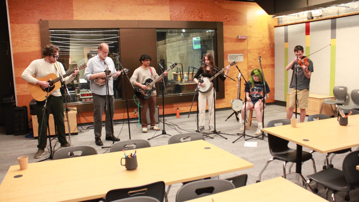 An adult playing an acoustic guitar, an adult playing a mandolin, a young adult playing a bass guitar, a young adult playing an banjo, a young adult sitting behind a microphone and playing spoons, and a young adult playing a violin into a microphone
