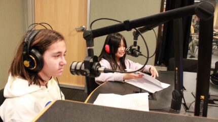 Two youth wearing headphones sitting behind microphones and pieces of paper in a radio studio