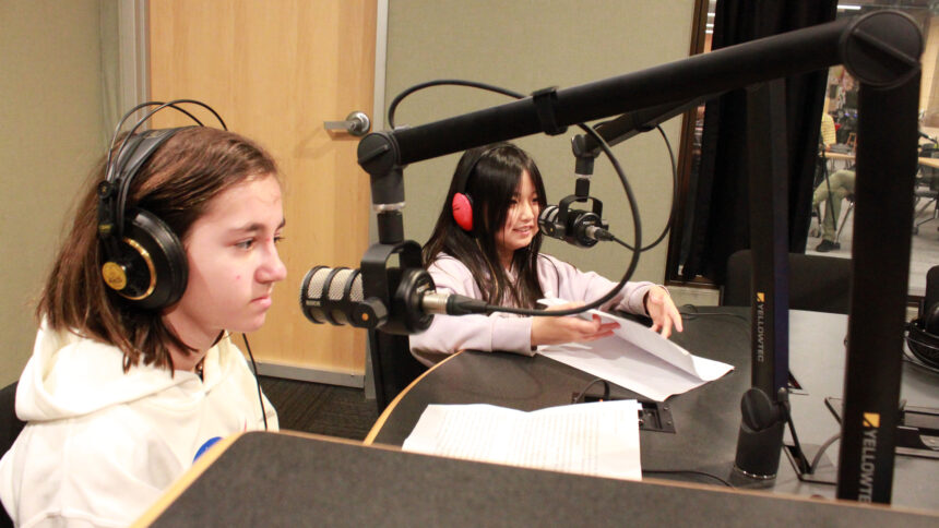Two youth wearing headphones sitting behind microphones and pieces of paper in a radio studio