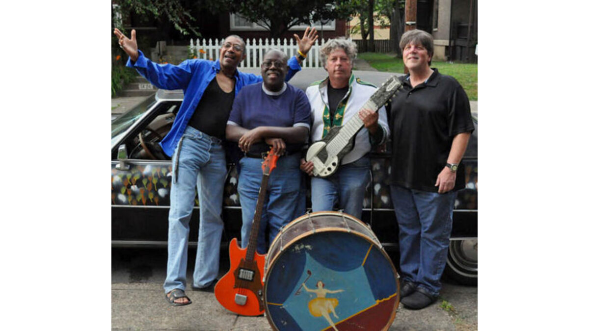Four adults posing in front of a car and behind a bass drum, one adult holding an electric bass, one holding an electric guitar