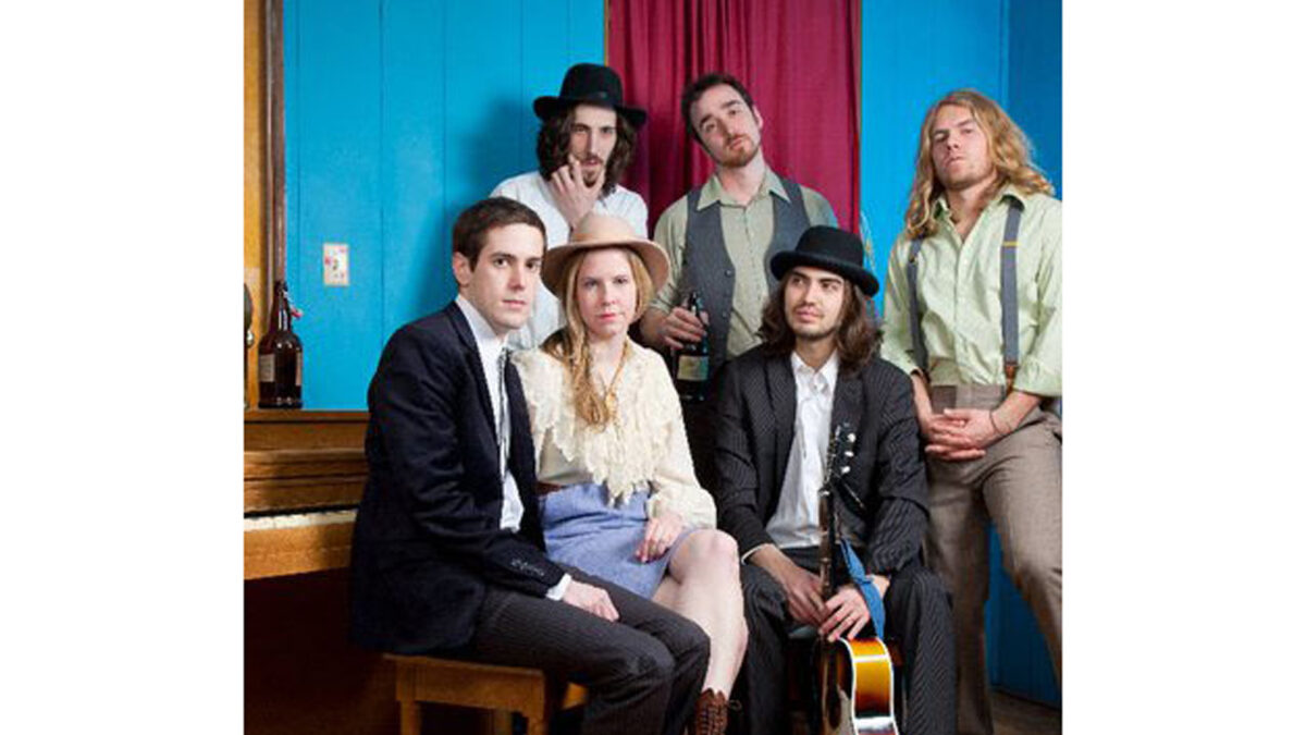 Six adults posing around a brown piano in a blue room