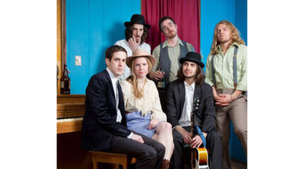 Six adults posing around a brown piano in a blue room