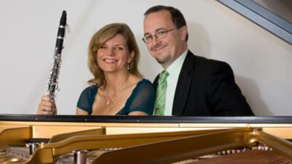 Two adults sitting behind a piano, one holding a clarinet