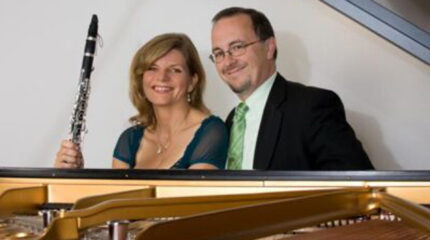 Two adults sitting behind a piano, one holding a clarinet