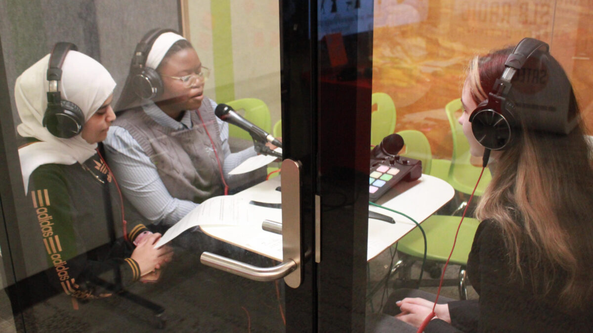 Three teens wearing headphones speaking into microphones in a recording pod