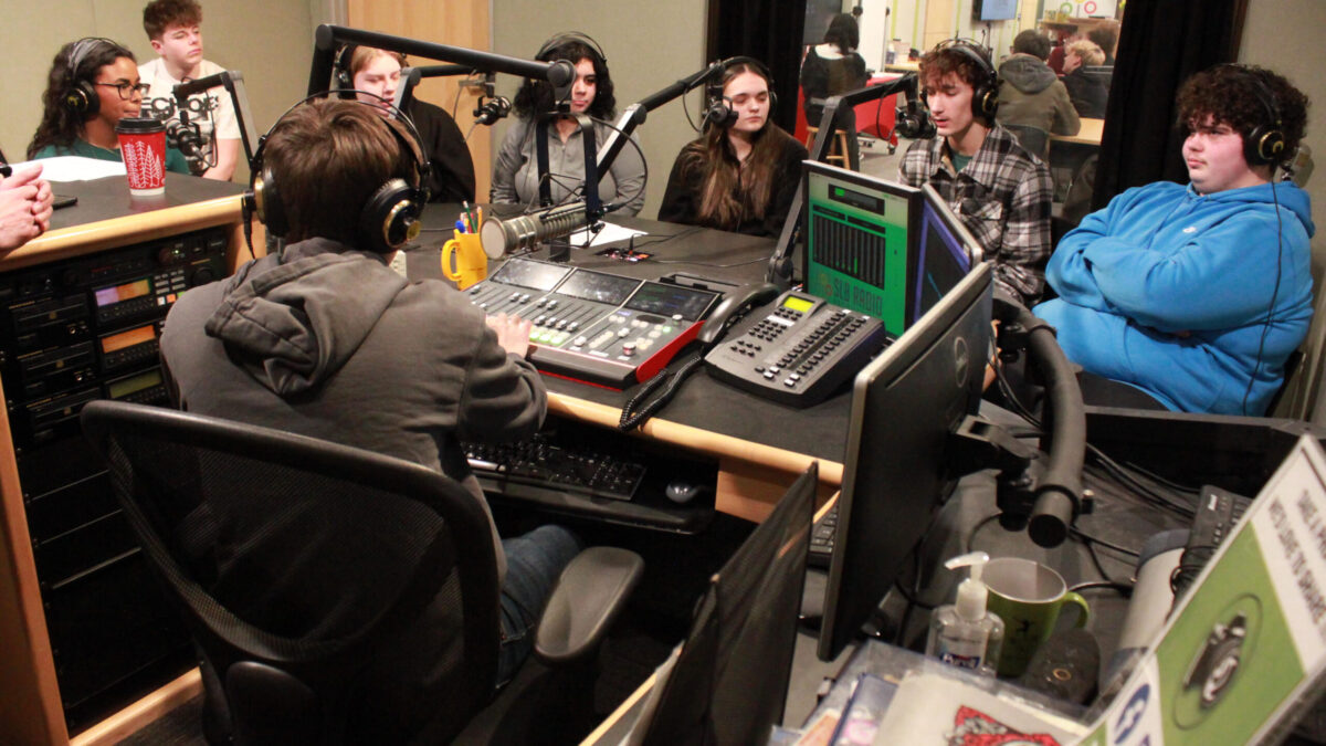 Eight teens wearing headphones sitting around microphones on a table in a radio studio