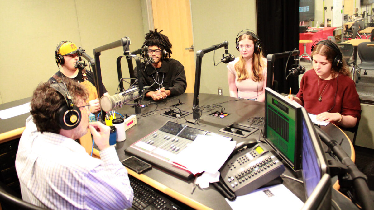 Four teens wearing headphones sitting behind microphones being interviewed by an adult wearing headphones sitting behind a microphone and an audio mixer in a radio studio