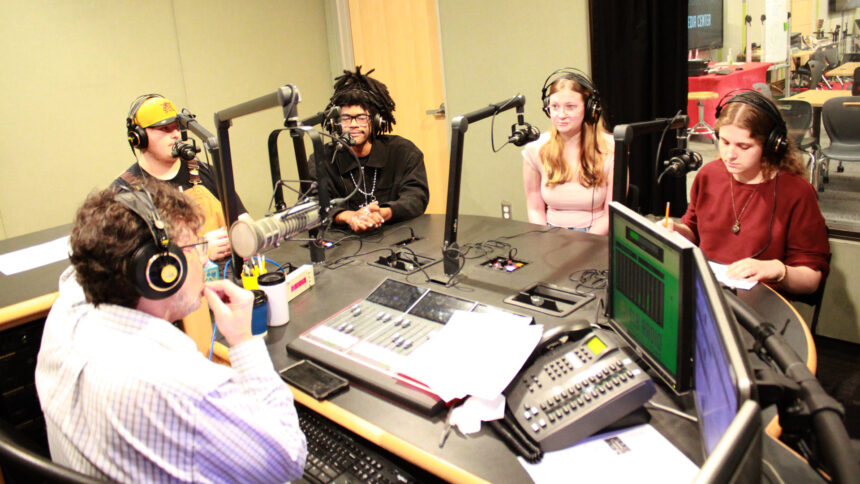 Four teens wearing headphones sitting behind microphones being interviewed by an adult wearing headphones sitting behind a microphone and an audio mixer in a radio studio
