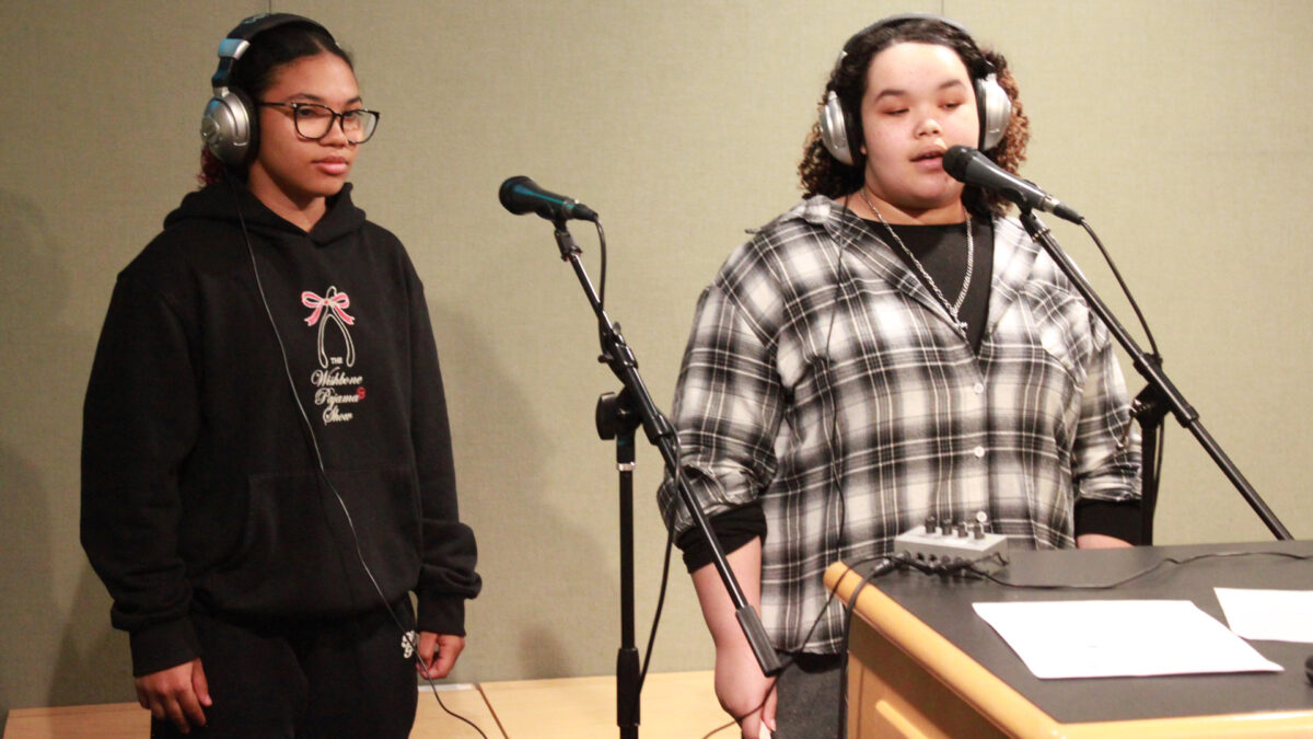 Two teens wearing headphones singing into microphones in a radio studio