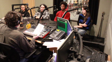 Four teens wearing headphones sitting behind microphones being interviewed by an adult wearing headphones and sitting behind a microphone and an audio mixer in a radio studio