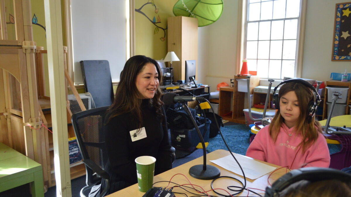two kids at a table wearing headphones listening to an adult sitting in front of a microphone