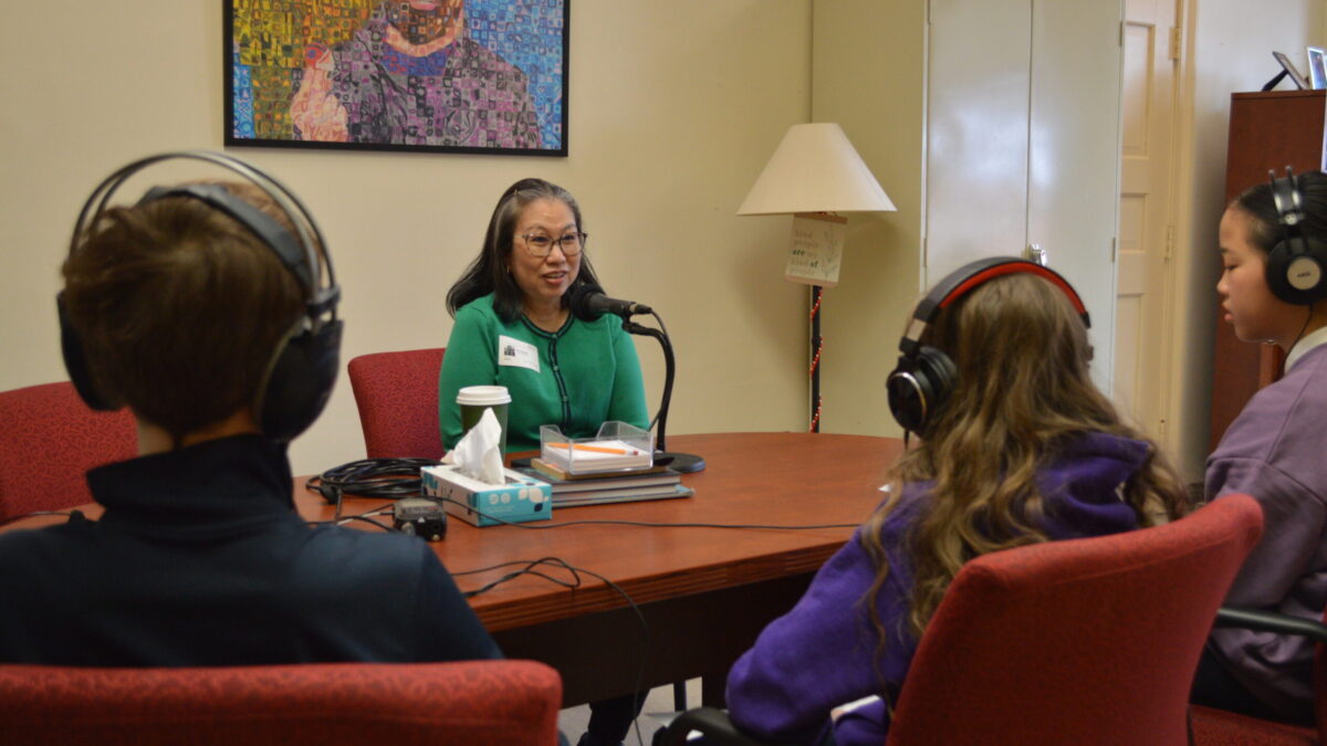 three kids at a table wearing headphones listening to an adult sitting in front of a microphone