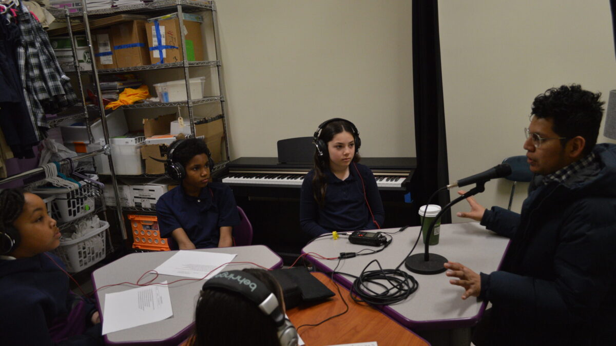 four kids at a table wearing headphones listening to an adult sitting in front of a microphone