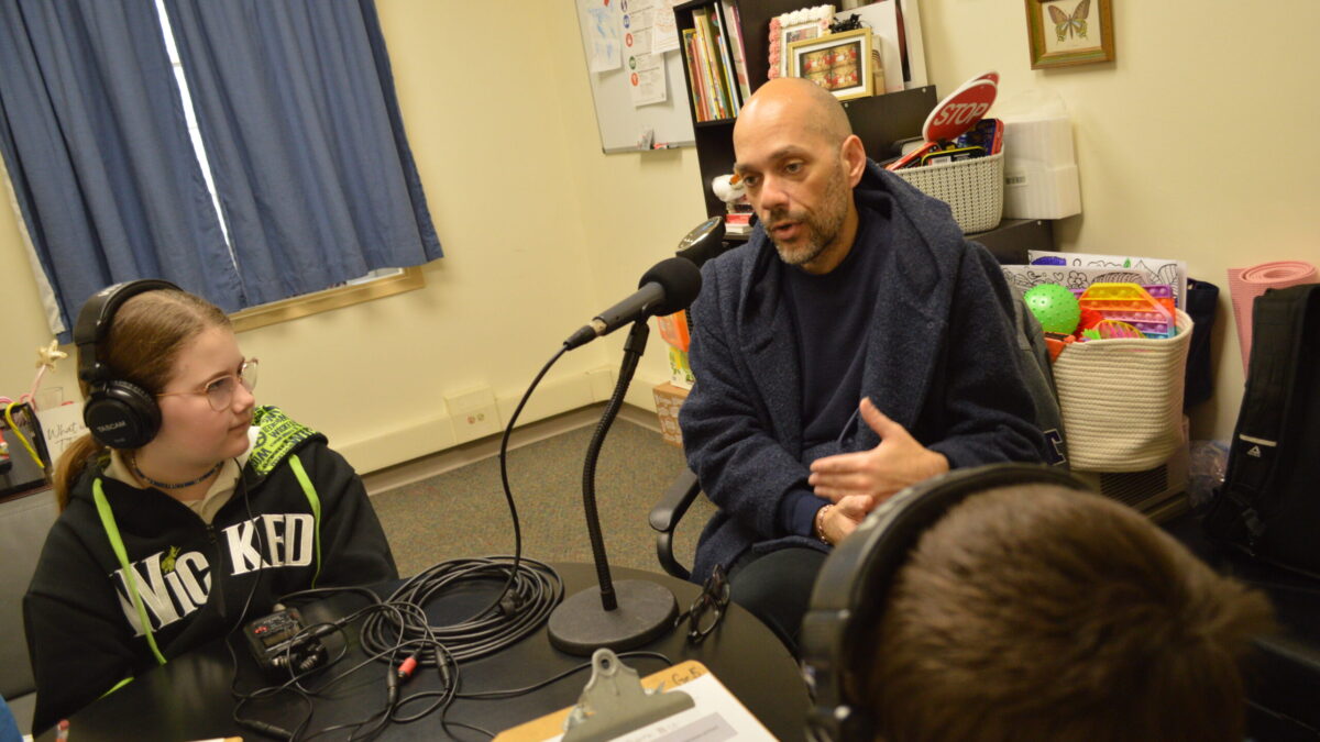 two kids at a table wearing headphones listening to an adult sitting in front of a microphone