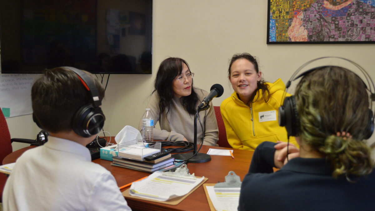 two kids at a table wearing headphones listening to two adults sitting in front of a microphone
