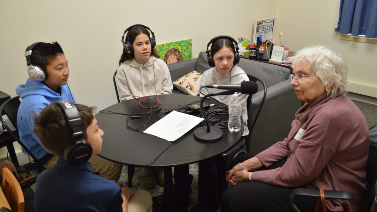 four kids at a table wearing headphones listening to an adult sitting in front of a microphone
