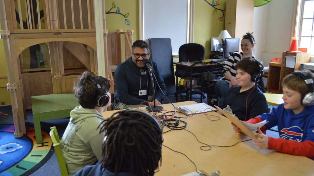 four kids at a table wearing headphones listening to an adult sitting in front of a microphone