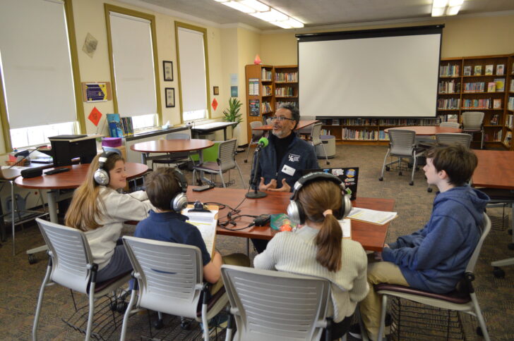 four kids at a table wearing headphones listening to an adult sitting in front of a microphone