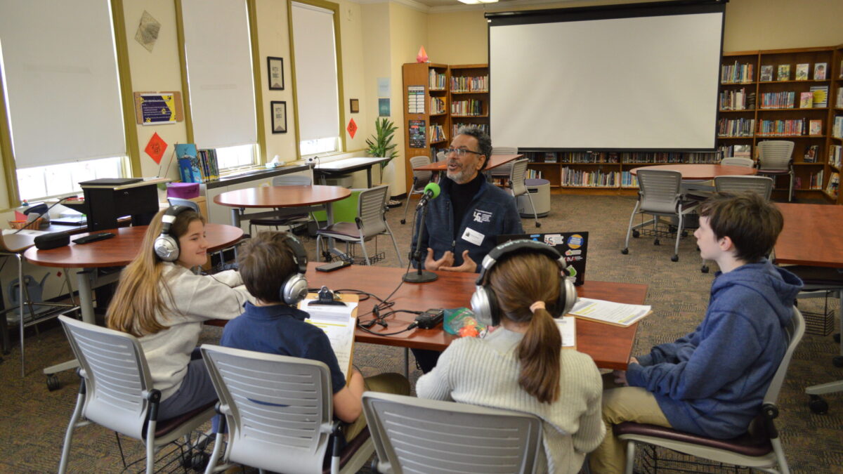 four kids at a table wearing headphones listening to an adult sitting in front of a microphone