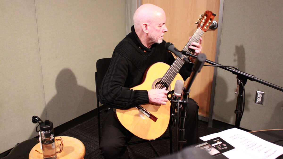An adult playing an acoustic guitar sitting behind microphones in a radio studio