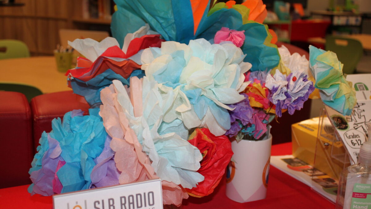 Two sets of paper flowers, one in a vase, on a red table