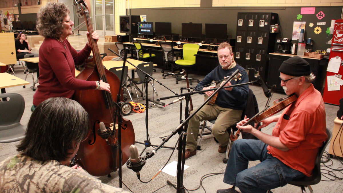An adult playing upright bass, an adult playing banjo, an adult playing fiddle, and an adult playing acoustic guitar