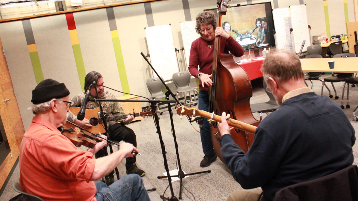 An adult playing fiddle, an adult playing acoustic guitar, an adult playing upright bass, and an adult playing banjo