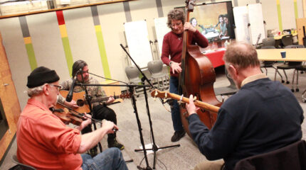 An adult playing fiddle, an adult playing acoustic guitar, an adult playing upright bass, and an adult playing banjo