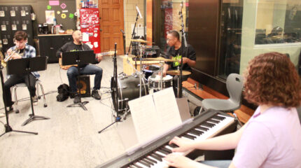 A teen playing trumpet, an adult playing upright bass, a teen playing a drum set, and a teen playing a piano keyboard