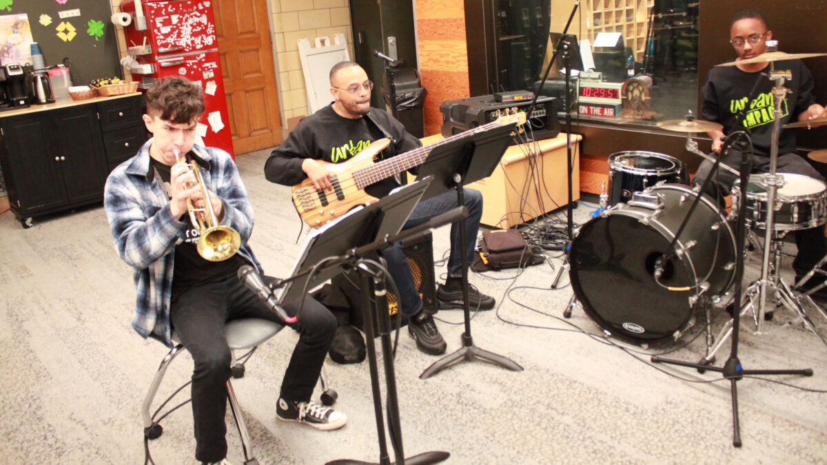 A teen playing a trumpet, an adult playing an electric bass, and a teen playing a drum set