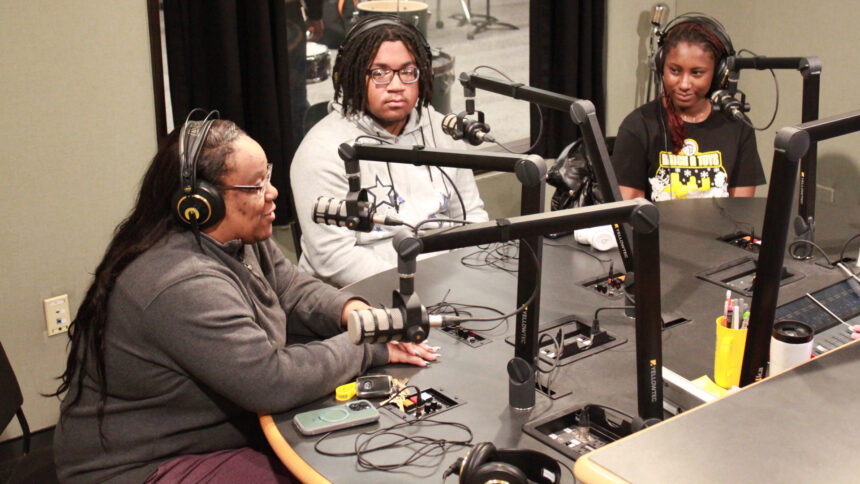 An adult and two teens wearing headphones speaking into microphones in a radio studio