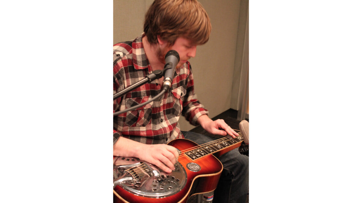 An adult playing a dobro sitting behind microphones in a radio studio