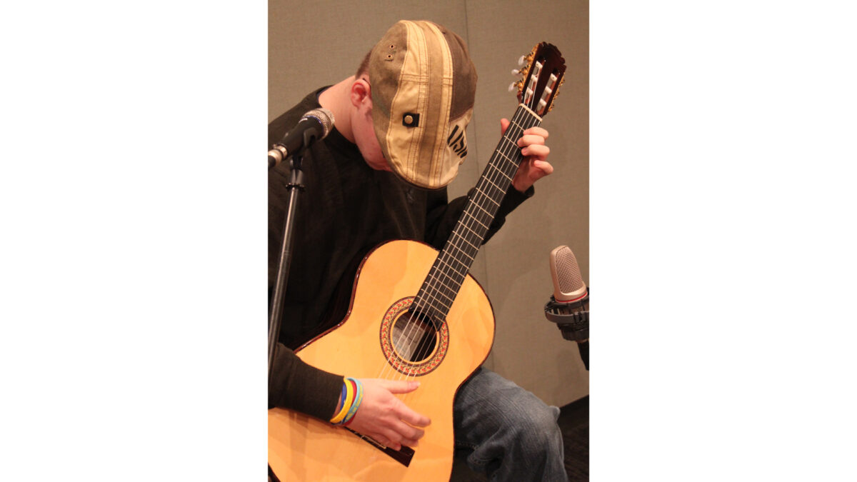 An adult playing an acoustic guitar sitting behind a microphone in a radio studio