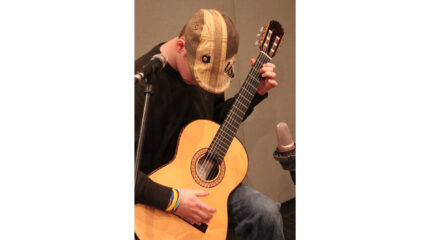 An adult playing an acoustic guitar sitting behind a microphone in a radio studio