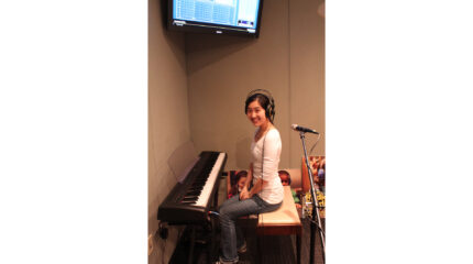 A teen wearing headphones sitting at a piano keyboard and smiling at the camera in a radio studio