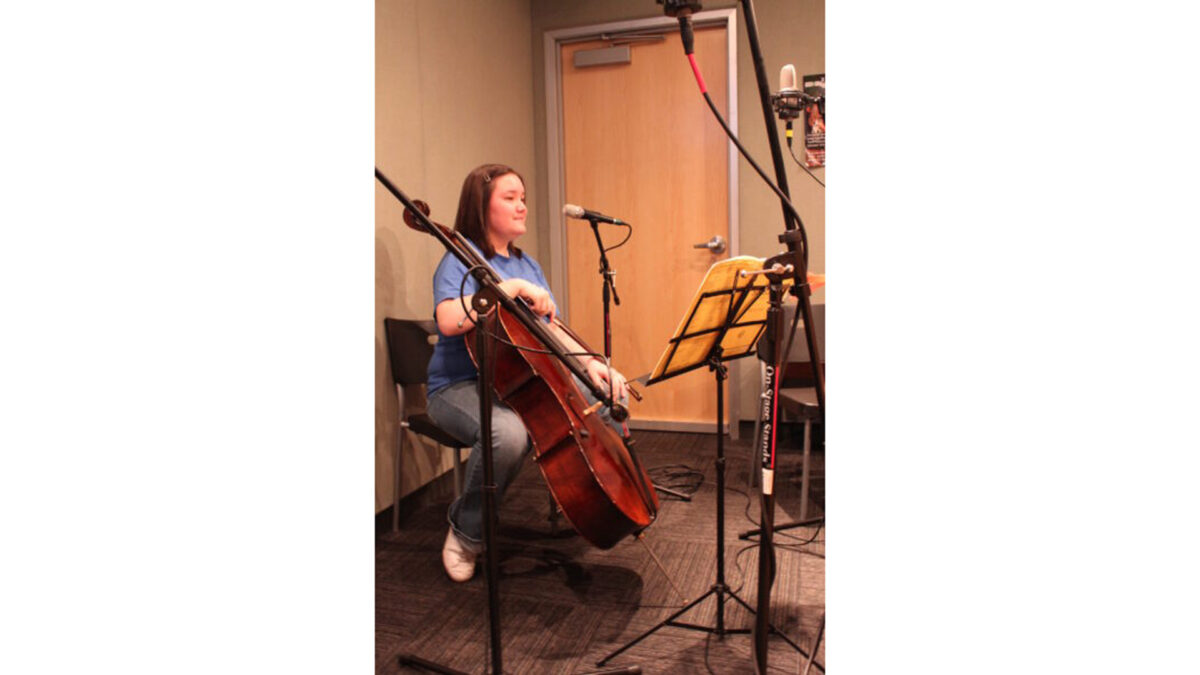 A teen holding a cello sitting behind a music stand with sheet music and two microphones in a radio studio