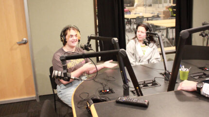 Two teens wearing headphones speaking into microphones in a radio studio
